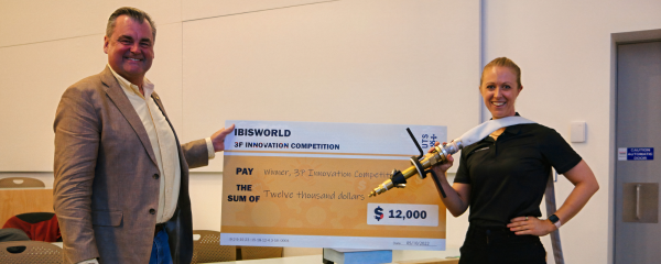 Sarah Battenally is presented with an oversize $12000 cheque. She holds her BAT product over her shoulder.
