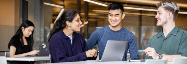 A female and two male IT students conversing around a laptop. One female student studies in the background