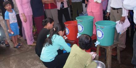 People crouching washing hands from buckets with taps and onlookers surrounding