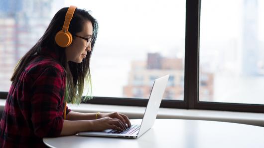 Female with orange headphones and glasses watching a laptop and typing a message. She is sitting in front of a large window overlooking apartments.