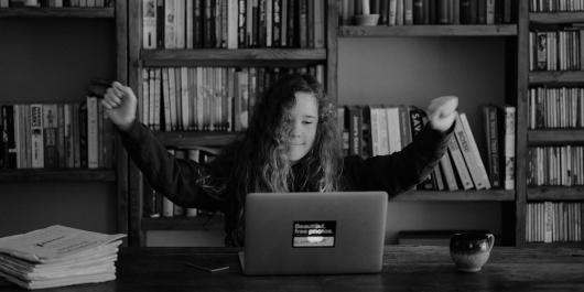 Black and white image of young girl with arms in air on laptop.