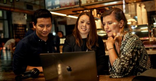 A man and two women sit at a cafe table and look at a laptop screen.