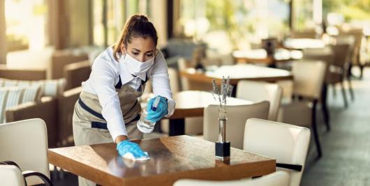 worker wearing a mask and gloves cleans a table in a restaurant