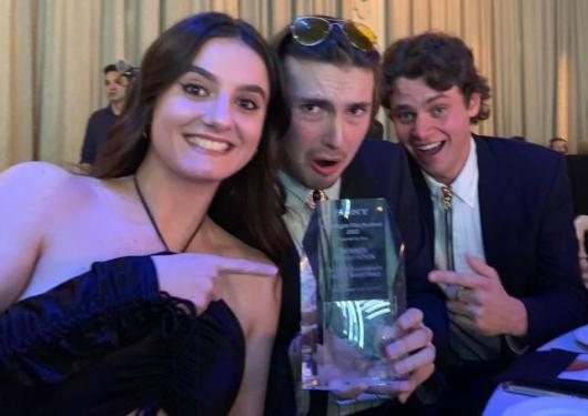 three students (one young woman, two young men) sitting together at a table wearing formal clothes, holding a trophy and smiling at camera
