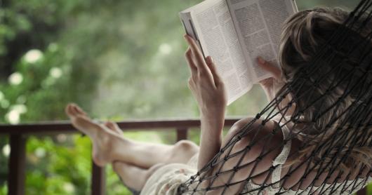 A person sits on a porch with her feet up and crossed while reading a book.