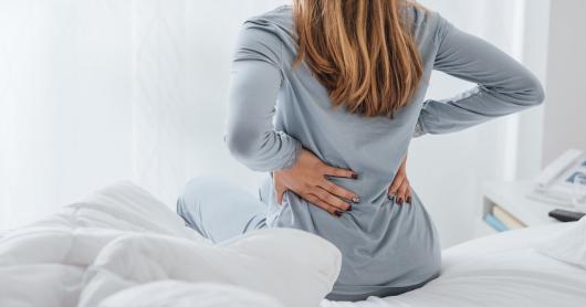 a person sits on a bed and clutches their back, seemingly in pain