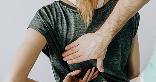 Clinician is examining a patient with back pain. The patient is a woman wearing a green t-shirt. She is sitting and the clinician's hands are on her back.