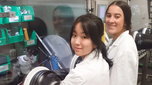 Two SQA Undergraduate Research Scholarship students in white lab coats looking over their shoulder and smiling.