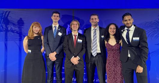 Six students - four male, two female - stand against a rich blue backdrop. They are wearing black suits and dresses, smiling. From left to right: Madeleine McWilliam; Cooper Crellin; Callum Burke; Matthew Dutaillis; Samantha Morris; and Sai Muthukumar