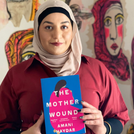 image of woman standing in front of artworks of women's faces. The woman is wearing a dark red shirt, headscarf and is holding a copy of a book. the book cover is blue and pink with an illustration of a woman and the text reads 'The Mother Wound' and 'Amani Haydar'.