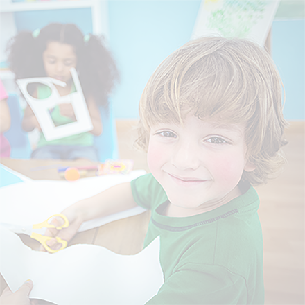 Happy kids doing craft. Blonde-haired boy looks at camera and smiling while cutting shapes in paper in foreground. Girl with dark curly hair in pigtails is cutting shapes from paper in the background.