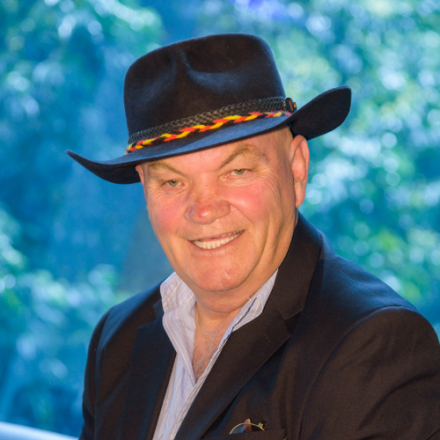 headshot image of Jack Beetson against a blue and green leafy background. Jack is wearing a wide brimmed black hat and smiling at the camera.
