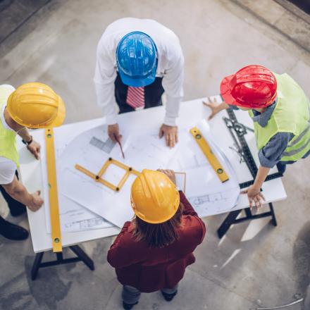 Four people in hard hats are discussing a plan. There are measuring instruments on the table and the photo is taken from above. 