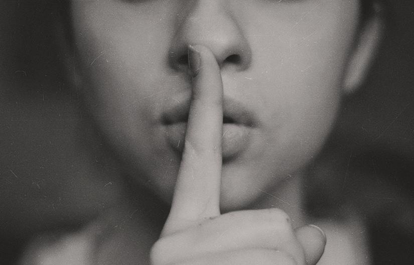 Close-up of a woman's face, with one finger making the 'shh' gesture in front of her lips