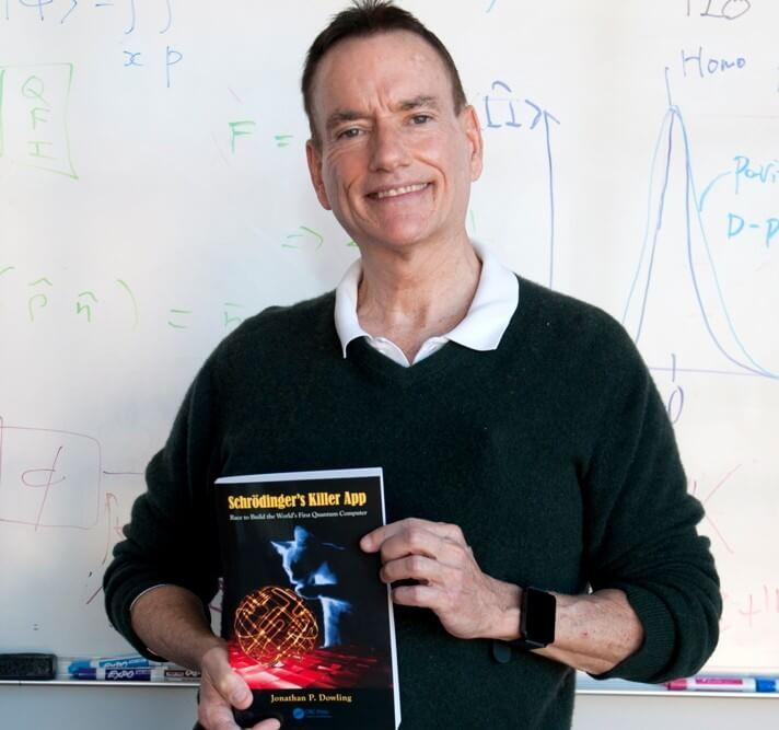 Photo of Prof Dowling in front a whiteboard covered in mathematical equations.  He is holding one of his books, Shrodinger's Killer App: Race to Build the World's First Quantum Computer