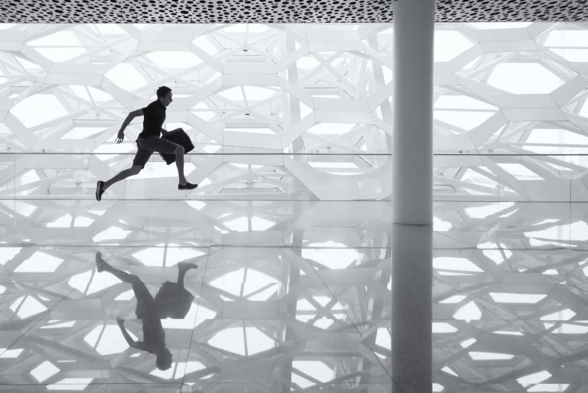 Man running with briefcase across a glass surface to reflect the geometric shapes of the building