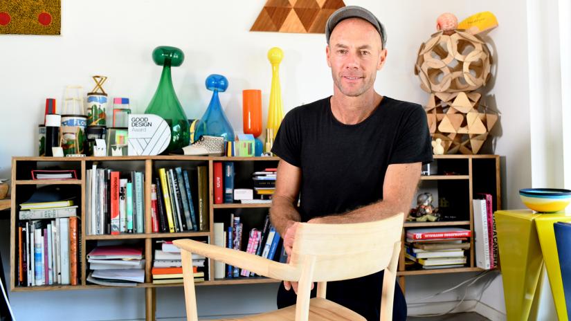 Adam Goodrum in his studio with his arm leaning on a library chair