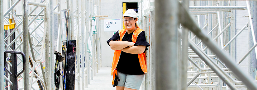 Young woman in hard hat and orange vest resting against metal pylons on construction site