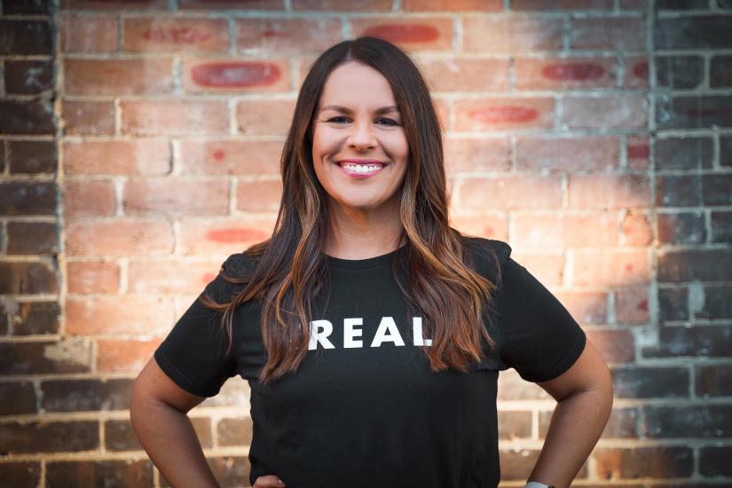 Image of women with long dark hair smiling at the camera. She wears a black t-shirt with the word REAL written across it in white. She is standing in front of a brick wall. She has her hands on her hips.