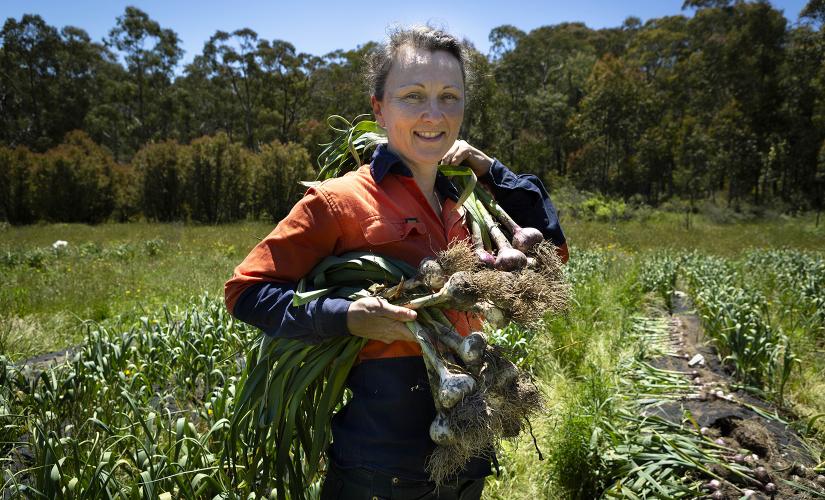 A woman in a high-vis orange shirt smiles at the camera and holds garlic plants, in front of a field filled with rows of garlic plants
