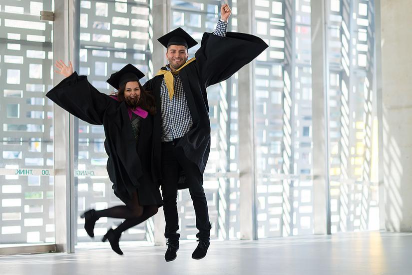 A man and woman in graduate academic gowns jumping happily in the air