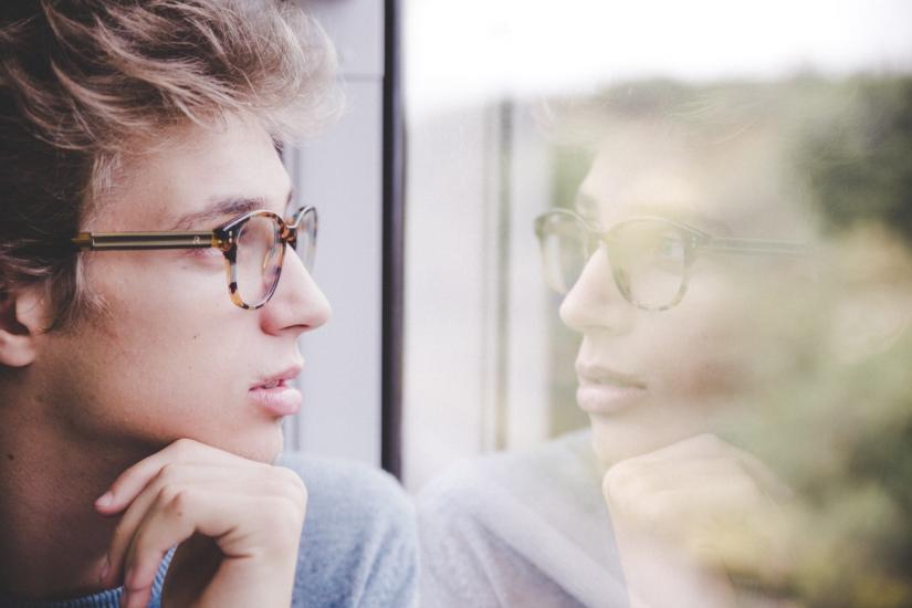 Young man with glasses looking at reflection in window