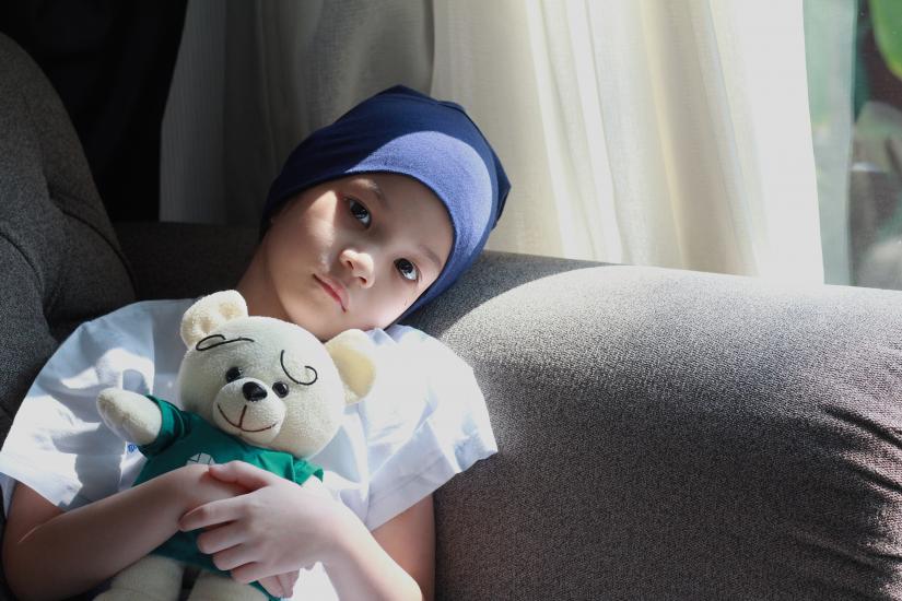 Child wearing head scarf, sitting on couch, holding teddy bear