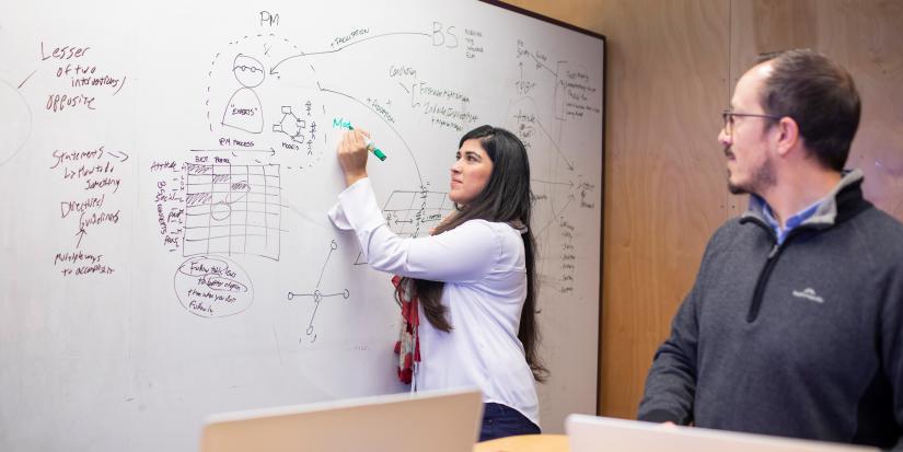 A man looks on as a woman solves the problem on a whiteboard