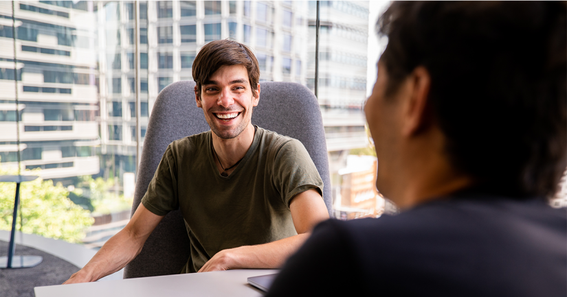 QSI's PhD student Fabio Henriques is sitting at a desk in conversation and laughing. In the background is a large window overlooking multistorey buildings opposite UTS Central on Broadway.
