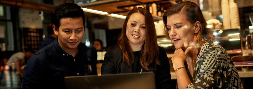 A man and two women sit at a cafe table and look at a laptop screen.