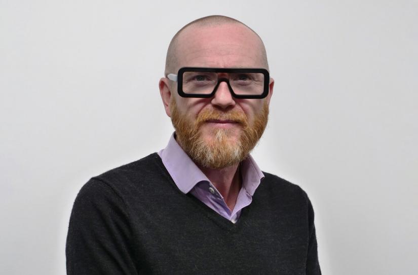 Chest up image of Director Jon Adams with thick black-framed glasses looking at the camera