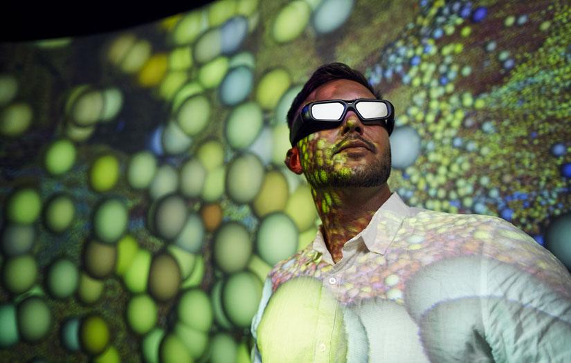 Young man looking off into the distance with 3D glasses on in front of a screen with green spheres