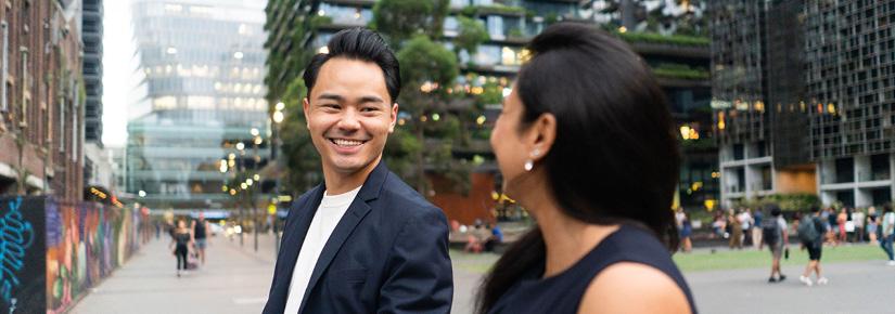 Two alumni walk through Central Park precinct, talking and smiling with greenery-covered towers in the background.