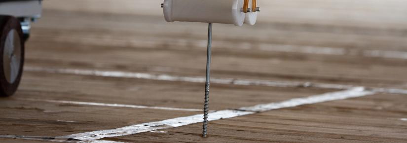 A robot arm screws a screw into a wooden floor.