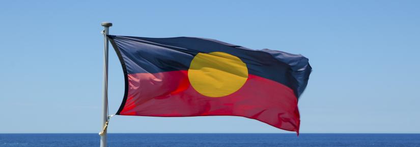 Australian aboriginal flag waving with blue sky and ocean water at the background.