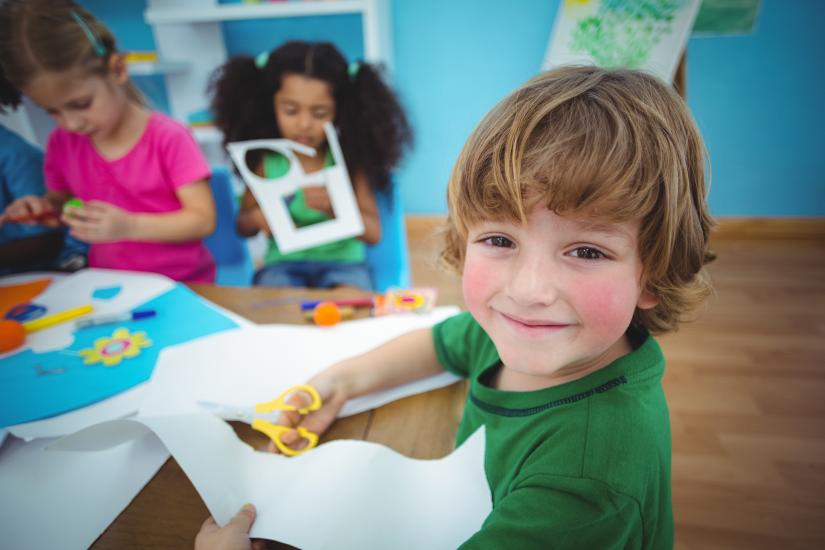 Kindergarten scene with children cutting shapes from paper. Blonde boy in foreground wears a green t-shirt and is smiling at thee camera. Children background are focus on cutting task.