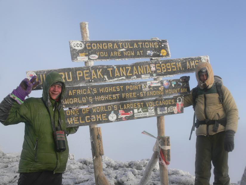 Two men stand on either side of a sign which reads "Congratulations you are now at Africa's highest peak"