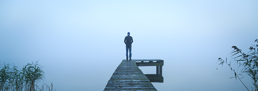 A man stands at the end of a jetty. The body of water sits under fog. Some water plants are visible on the left and right of the image. 