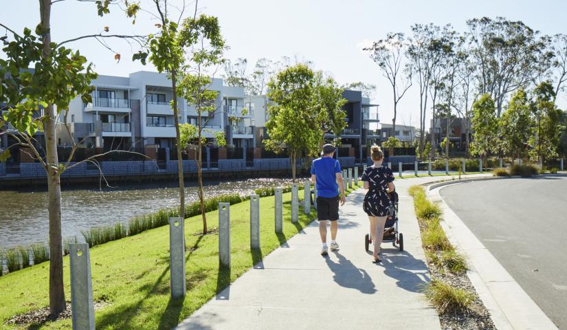 Two people walk along a pathway, a road on one side, and a waterway and buildings on the other