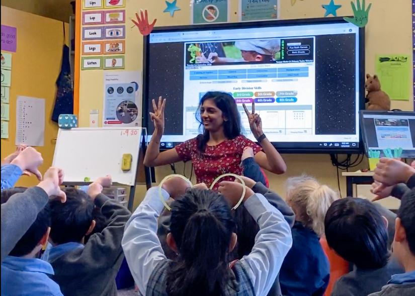 A female teaching student holds her hands up to a classroom of primary children