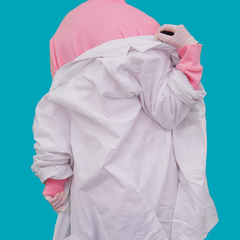 A person viewed from the back removing a lab coat to show a pink shirt underneath