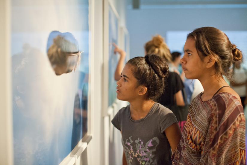 Two young Indigenous women in an art gallery observing a photographic print on the wall. One of the women is leaning in for a closer look. The photograph is of a feather against the backdrop of a sunny blue sky. It is by Wiradjuri/Kamilaroi artist Michael Riley, and the name of the work is Untitled (Feather) 2000.