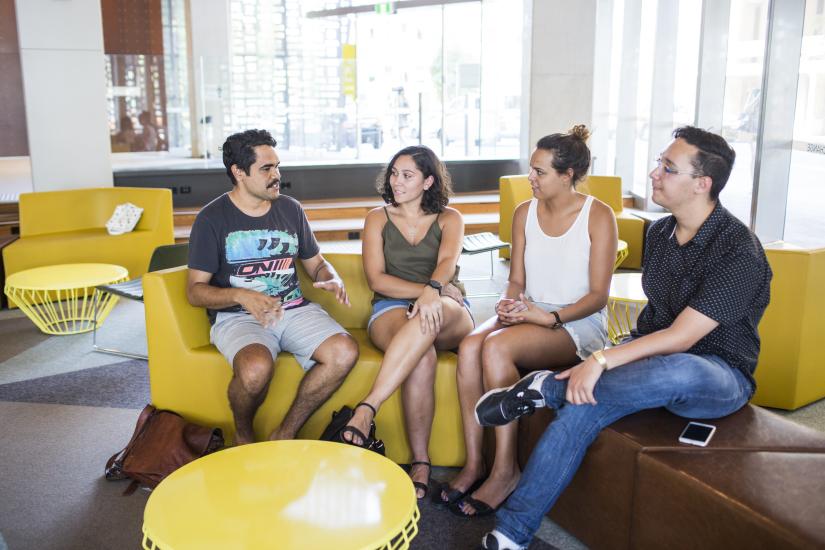 A group of Indigenous students, two male and two female, are sitting on a couch engaging in casual conversation.