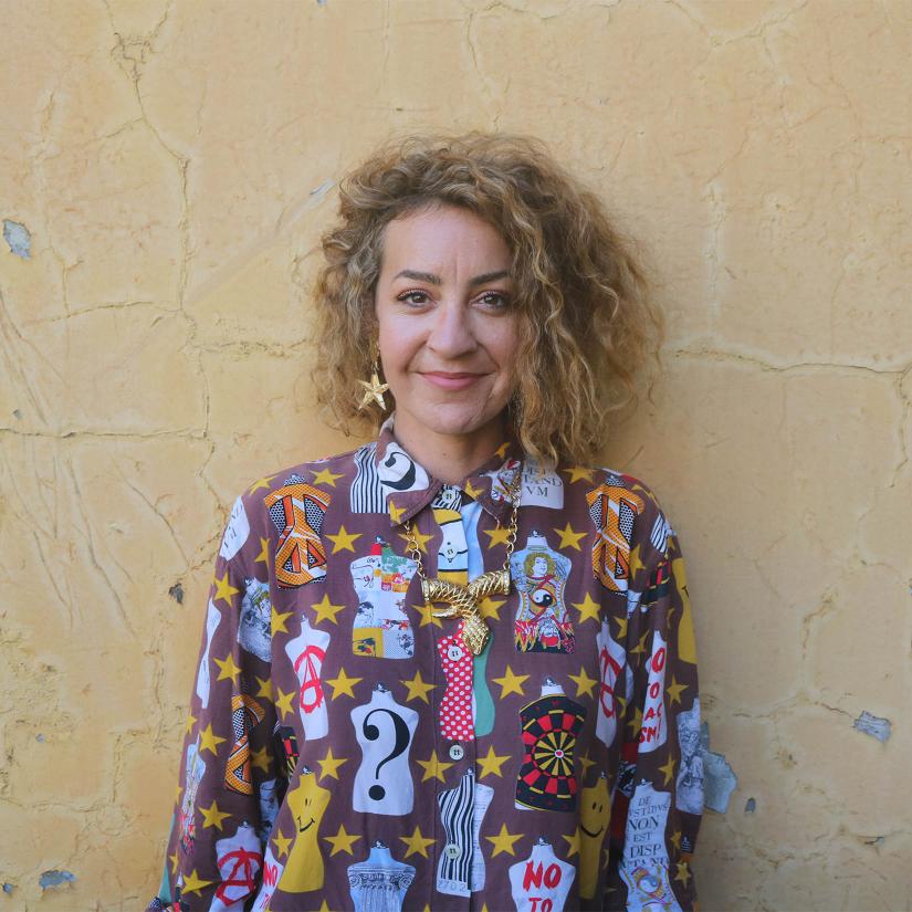 A smiing person in a colourful shirt stands against a textured yellow wall