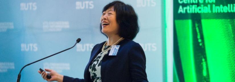 Distinguished Professor Jie Lu delivering a speech in front of a podium, smiling with her arms outstretched.