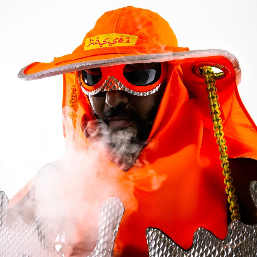 A figure wearing orange sunglasses and a hat blows smoke towards the camera