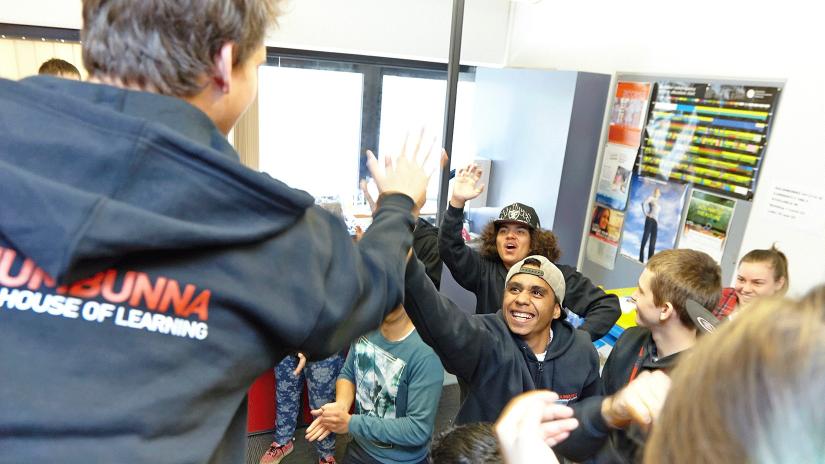 A group of Indigenous high school students cheerfully participating in a Galuwa program at Jumbunna. Two smiling students in the middle of the group reach out to give a high-five to an encouraging program instructor.