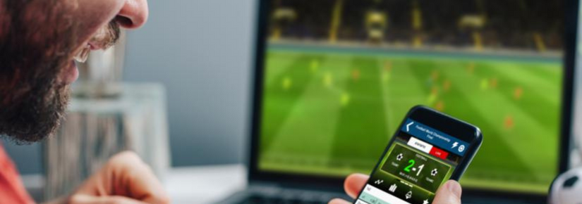Stock photograph of man gambling on mobile phone while sports play on laptop in background.
