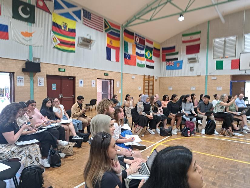 Image of 50 plus university students sitting on chairs in a semi-circle in a high school gym. There are coloured flags of the world on the walls behind them.