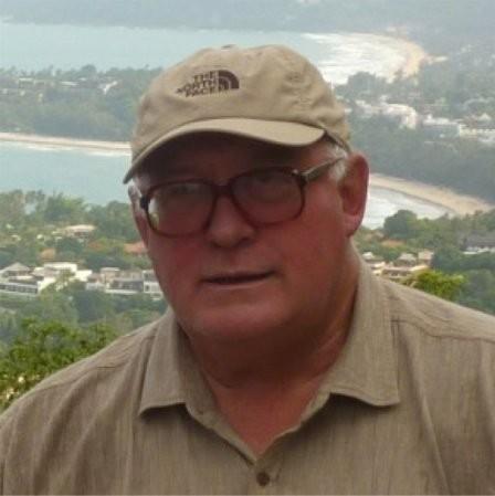 Square shot of Tim Hassett in baseball cap and glasses, head and shoulders visible against a residential coastal background